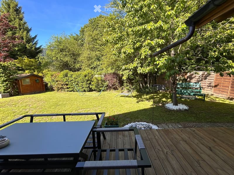Well-maintained garden with a wooden deck, outdoor table and chairs, small shed, and a green bench in the background.