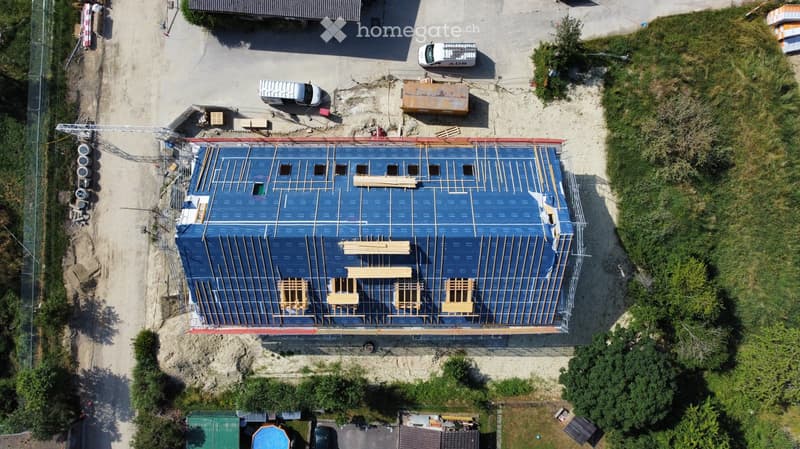 Aerial view of a building under construction with a blue tarp-covered roof and exposed wooden beams. Surrounding area includes construction materials, vehicles, and greenery.