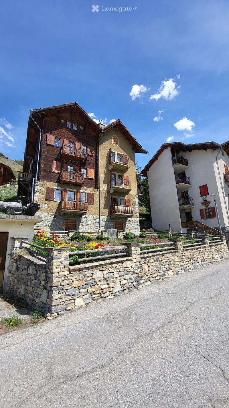 A multi-story residential building with wooden balconies and stone facades, surrounded by a stone wall and colorful flowers.