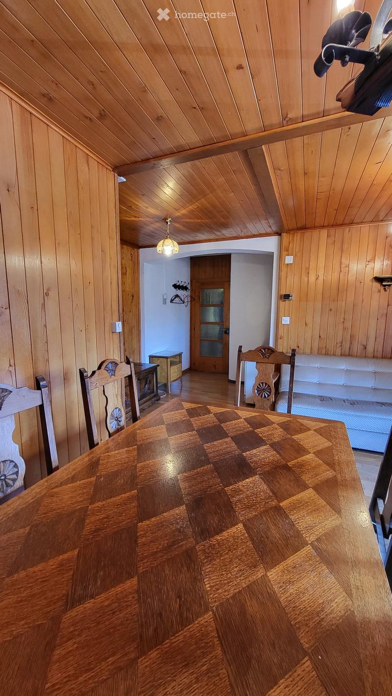 A cozy, wooden-paneled room with a checkered wooden table, chairs, a bed, a nightstand, and a fireplace in the background.