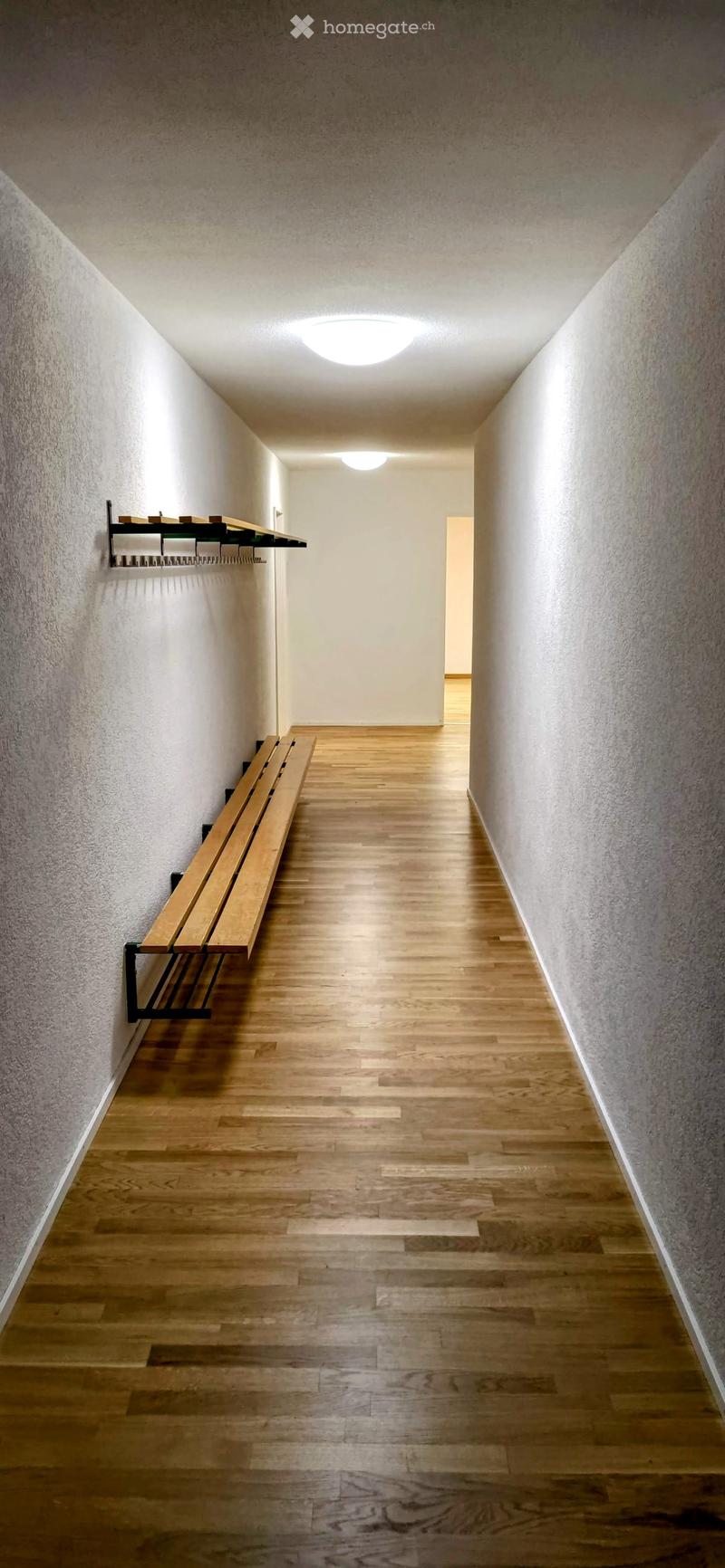 A modern hallway with wooden flooring, white walls, and built-in wooden benches on one side. The ceiling features recessed lighting.