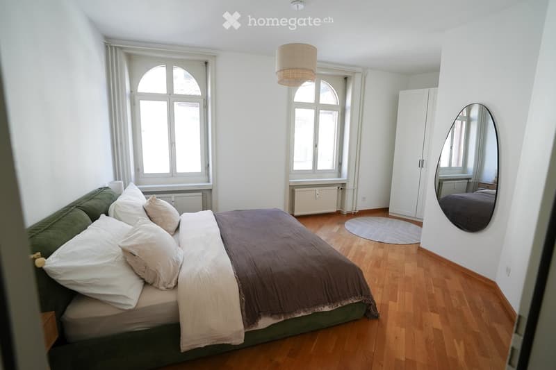 Cozy bedroom with a wooden floor, large windows, a bed with pillows and a blanket, a green sofa, a round rug, a large mirror, and a wardrobe.