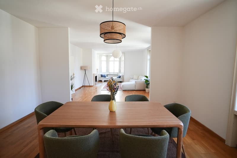 Bright, open living and dining area with wooden flooring, large windows, a dining table with six chairs, a sofa, an ottoman, a floor lamp, and decorative vases.