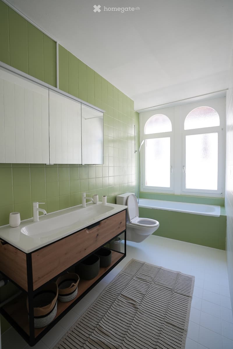 Modern bathroom with green tiled walls, white sink with wooden cabinet, toilet, bathtub, and large windows allowing natural light.