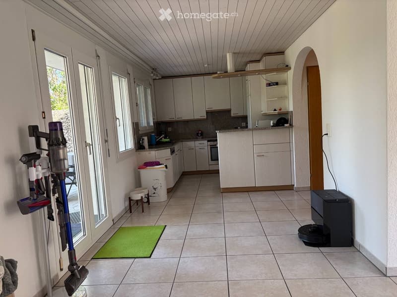 Bright, tiled entryway leading to a modern kitchen with white cabinets, stainless steel appliances, and a countertop. A vacuum cleaner is mounted on the wall, a pet bowl and stool are near the trash bin, and a green rug lies on the floor.