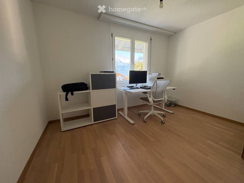 A modern, well-lit room with wooden flooring, a white desk, a chair, a screen, a keyboard, and a shelf with a bag on it.