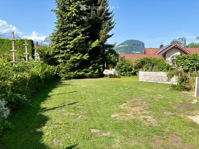A well-maintained garden with a lawn, tall trees, a small patio area with a table and chairs, and a wooden fence. The background features a house with a red-tiled roof and distant mountain views.