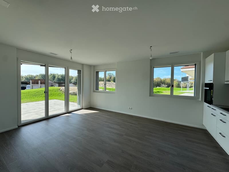 Empty room with light grey walls, wooden flooring, large windows, and sliding glass doors leading to a patio.