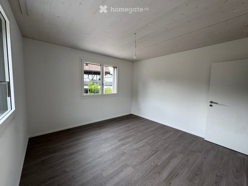 Empty room with light-colored walls, wooden flooring, and a single window showing a view of the outside. The room has a simple, modern design with a door on one side.