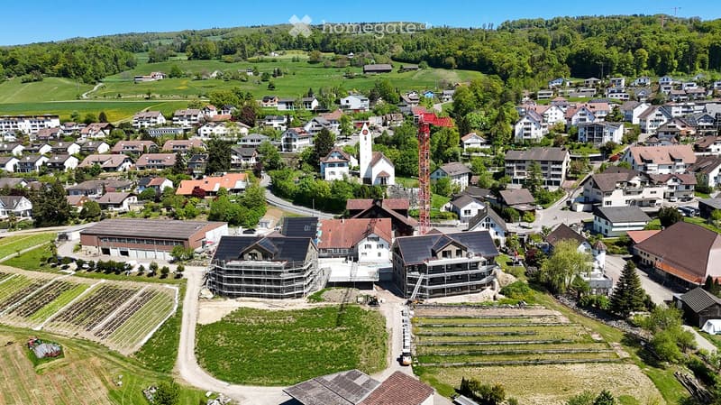 Aerial view of a small village with numerous houses, a church tower, and a construction site with scaffolding and cranes.