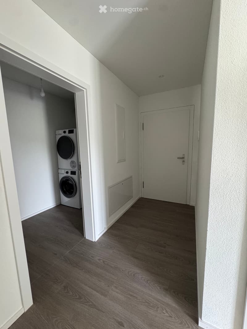 Empty hallway with wooden flooring, white walls, and a washing machine in an adjacent room.