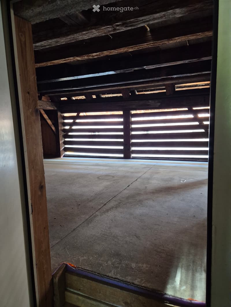Empty room with concrete floor, wooden ceiling beams, and a window with horizontal slats allowing natural light to enter.