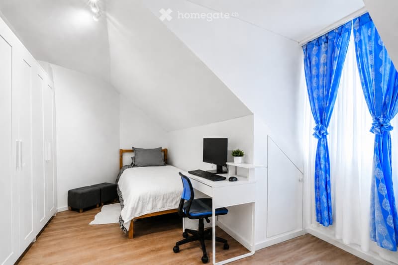 Cozy attic bedroom with a bed, desk, chair, computer setup, and small plant. Features wooden flooring, white walls, and blue curtains.