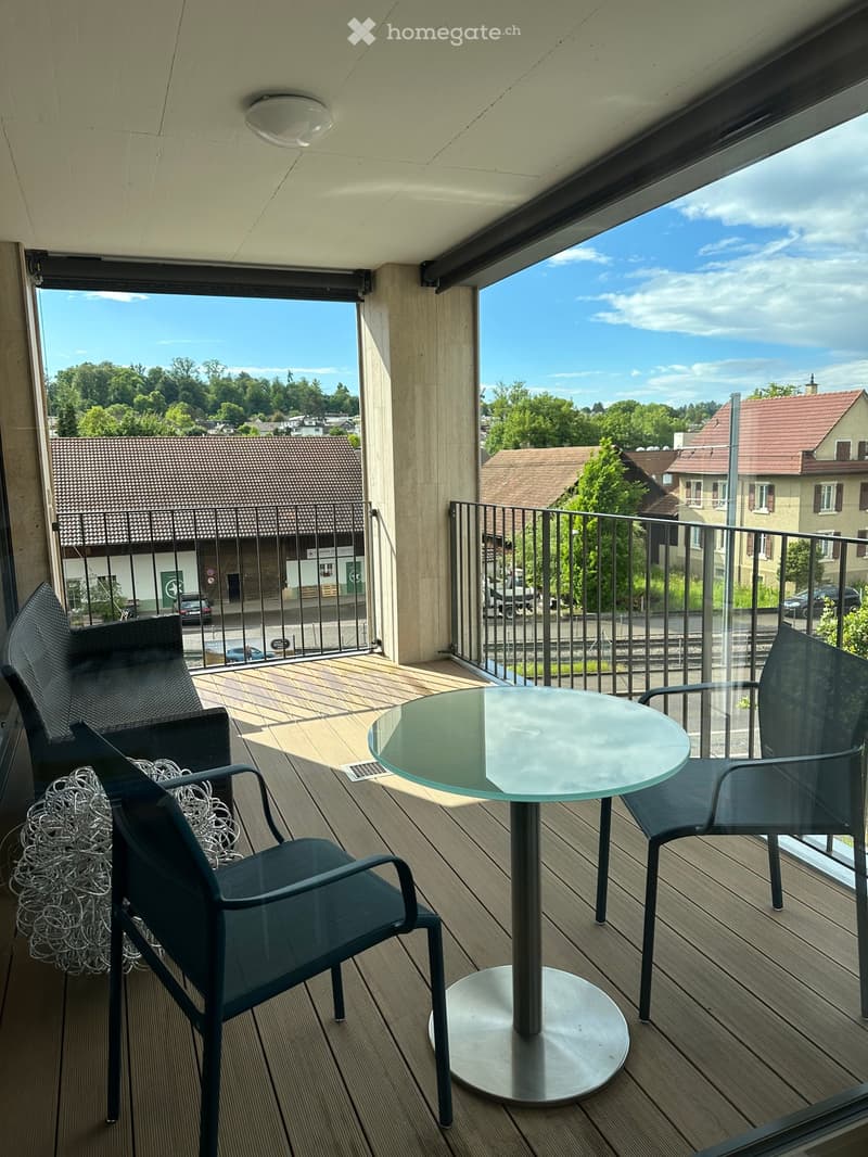 Balcony with wooden decking, glass table, and two chairs, overlooking a town with houses and trees.