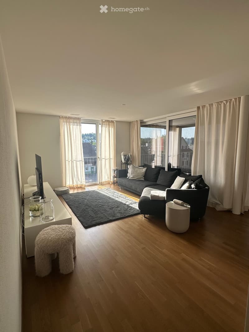 Modern living room with wooden flooring, large windows, grey sofa, black rug, and decorative items on a white console table.