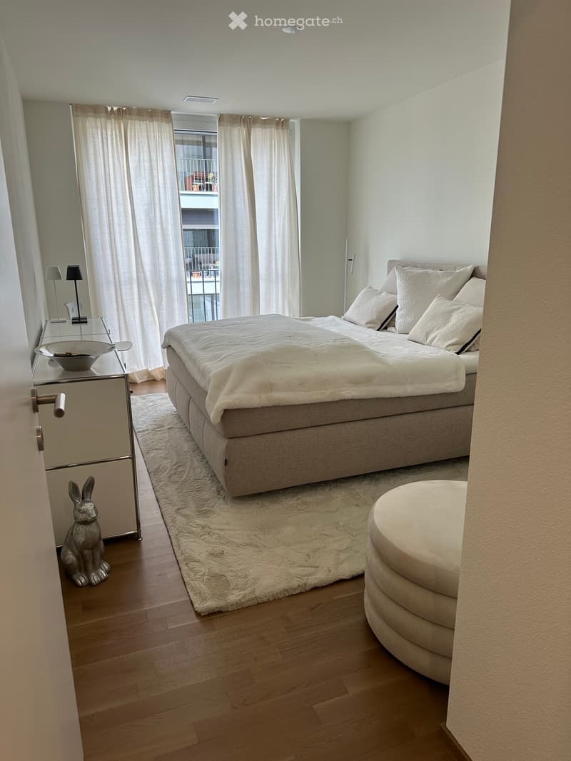 Modern bedroom with a large bed, nightstand, stool, and a view of a balcony through sheer curtains.