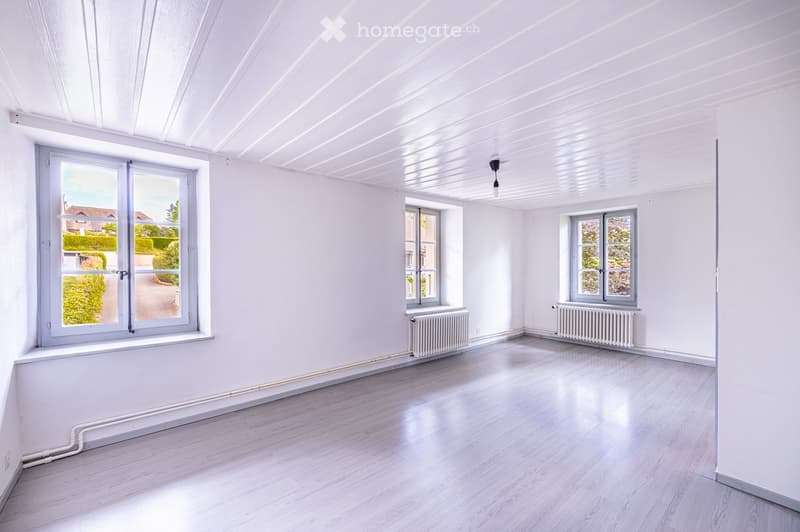 Empty room with white walls, light wooden flooring, and large windows allowing natural light. Radiators are present under the windows, and the ceiling has a modern, ribbed design.
