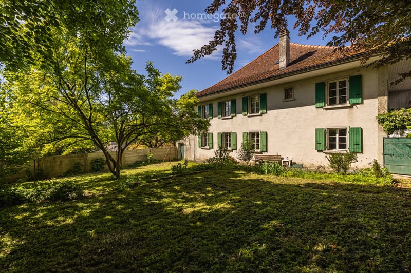 A two-story house with a red-tiled roof and green shutters, surrounded by a lush garden with trees and a bench.