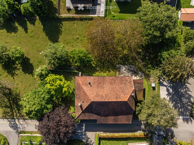 Aerial view of a two-story house with a brown tiled roof, surrounded by a well-maintained lawn and trees. The house has multiple chimneys and is situated near a road with neighboring houses visible in the background.