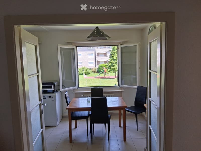 Bright kitchen with tiled floor, large window overlooking a green area, table with four chairs, dishwasher, and printer on a counter.