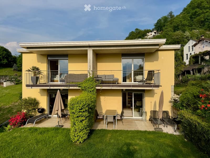 Two-story modern house with yellow facade, large windows, and balconies. Features include a well-maintained garden, patio seating, and outdoor dining area.