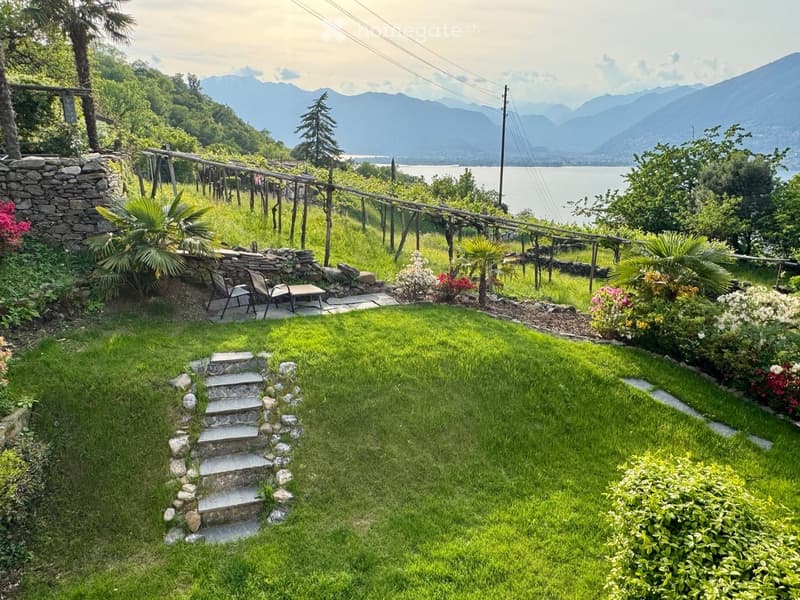 A well-maintained garden with green grass, stone steps, a small seating area with chairs and a table, surrounded by various plants and trees, with a scenic mountain and lake view in the background.