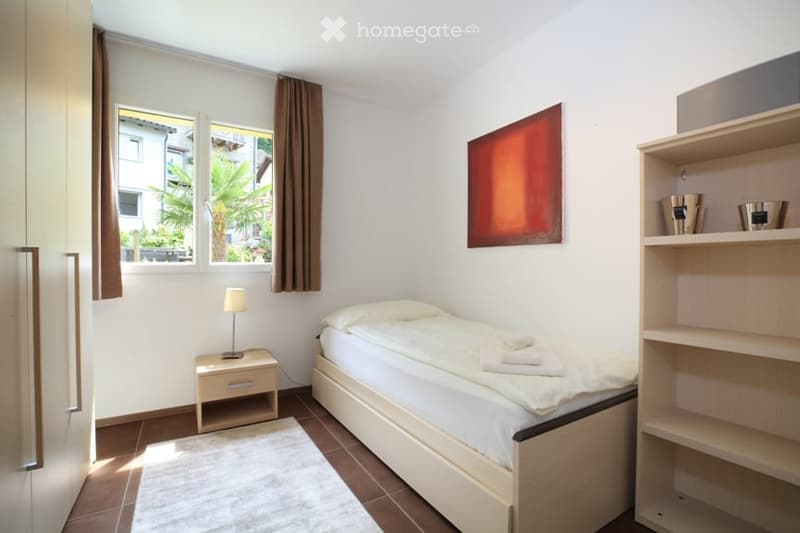 Cozy bedroom with a single bed, nightstand with lamp, wooden shelf, and a large window with brown curtains.