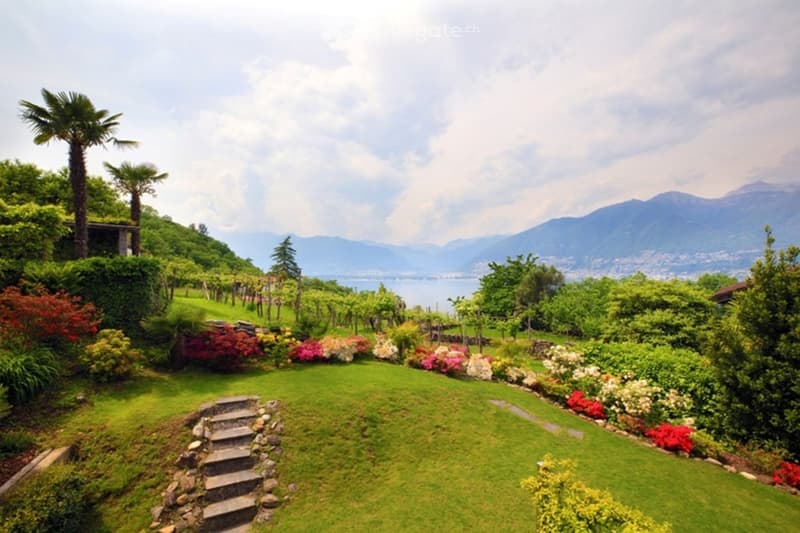 A lush garden with green lawns, colorful flowers, trees, and a scenic view of a lake and mountains in the background.