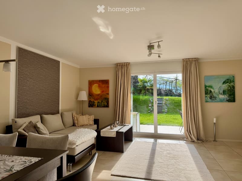 Bright living room with large sliding glass doors opening to a garden, beige sofa, coffee table, dining table, and artwork on the walls.