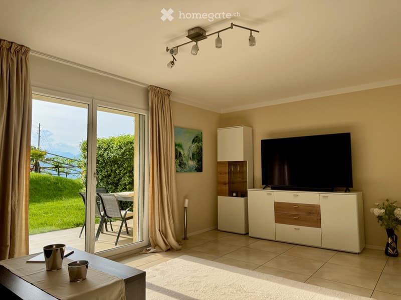 Bright living room with large sliding glass doors leading to a patio, a TV on a white cabinet, a small table with two silver cups, a painting on the wall, and a view of greenery and mountains.