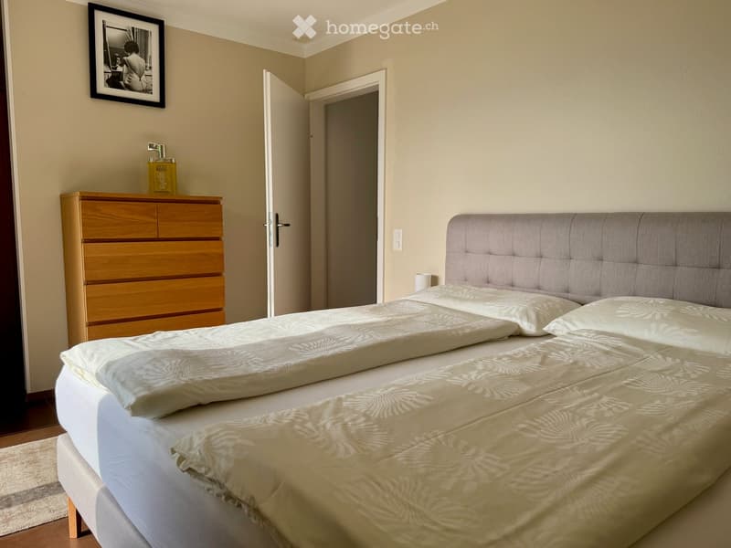 A well-lit bedroom with a large double bed, a wooden dresser, and a picture frame on the wall.