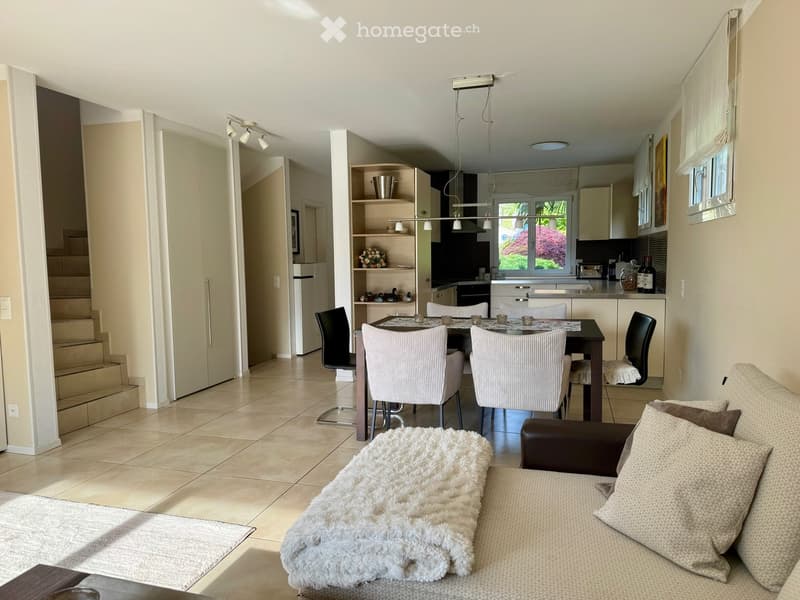 Modern living and dining area with a sofa, coffee table, dining table with chairs, and an open kitchen area with cabinets, appliances, and a window view.