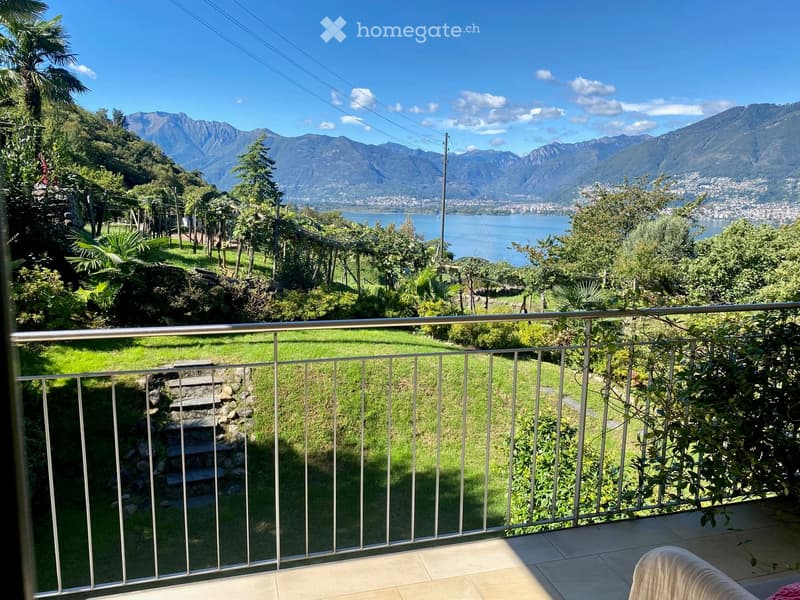 Balcony with metal railing overlooking a lush green garden, stone steps, and a scenic view of a lake and mountains.