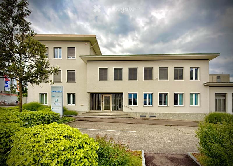 Modern two-story building with a flat roof, white facade, multiple windows, and a sign reading "BRODECK" on the left side. The entrance features a set of steps leading to a glass door.