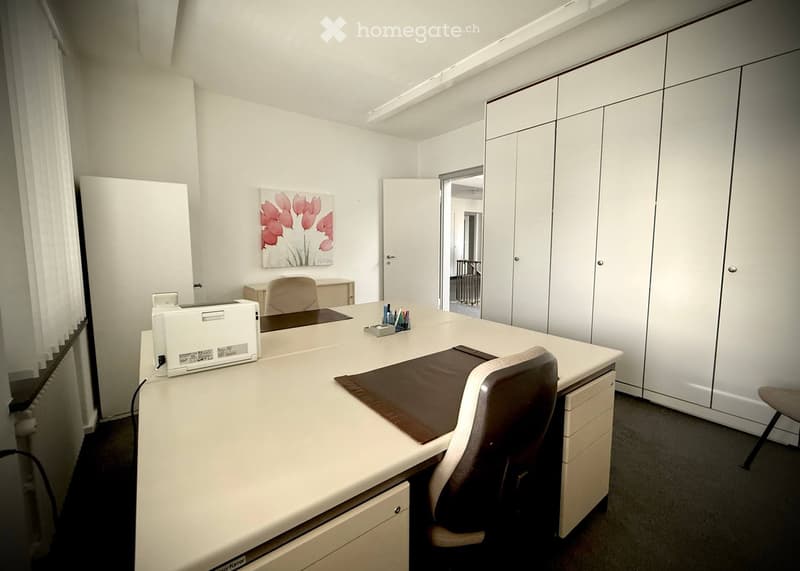 Office space with a white desk, ergonomic chair, printer, and built-in white cupboards. A picture frame with a floral design hangs on the wall.