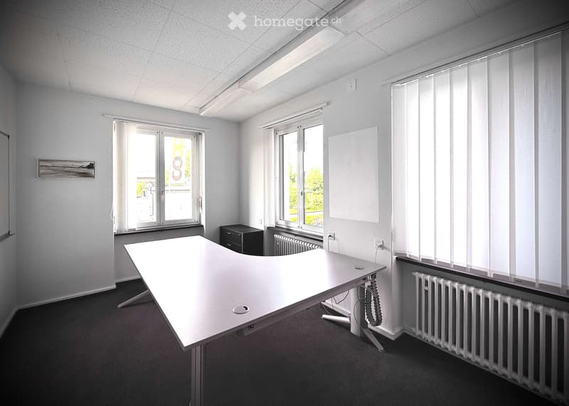 Modern office space with a large white desk, radiator, and cabinet. Large windows with blinds and a view of greenery.