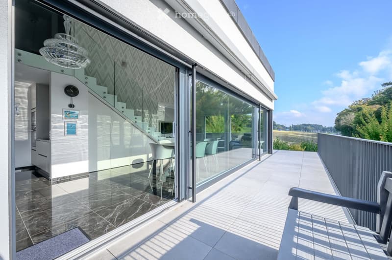 Balcony / Terrace. Modern balcony with glass walls, tiled floor, and metal railings. Features a chandelier, a table with chairs, and a bench. The interior reflects through the glass, showing a staircase and kitchen.