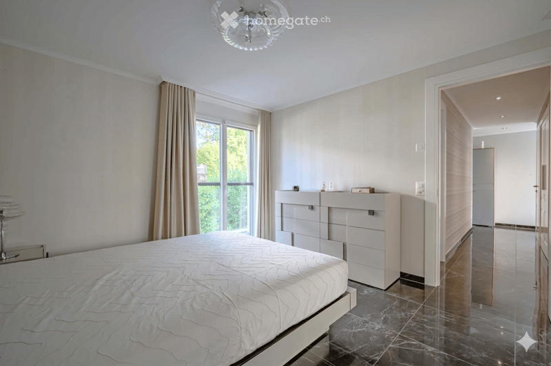 Bedroom. Spacious bedroom with a large bed, white walls, and marble flooring. A nightstand with a lamp is on the left, and a set of drawers is against the wall. The room has a large window with curtains.