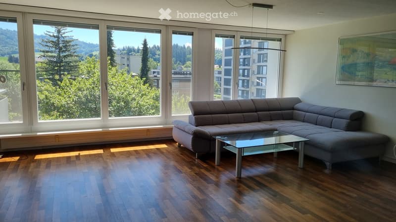 Modern living room with large windows offering a scenic view, featuring a grey sectional sofa and a glass coffee table on a wooden floor.