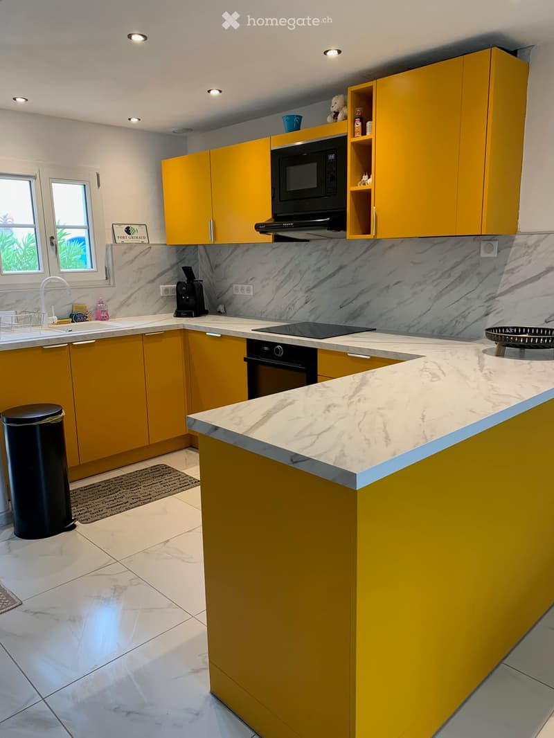 Modern kitchen with yellow cabinets, marble countertops, and stainless steel appliances including a microwave, oven, and coffee maker. Features a central island with a sink and faucet, and a window with a view of greenery.