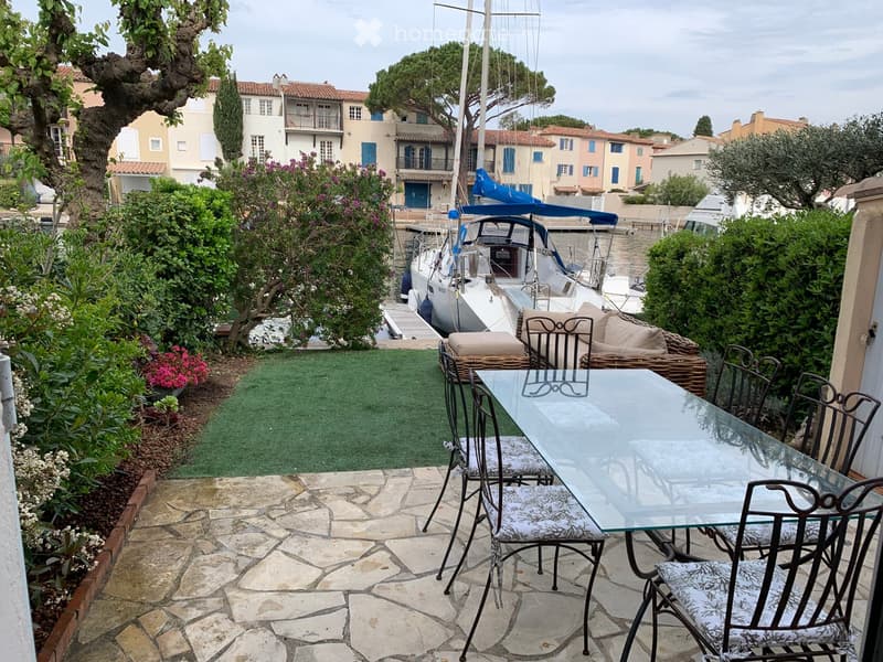 Outdoor patio with a glass table, chairs, and a small sofa, surrounded by greenery and a view of a marina with sailboats.