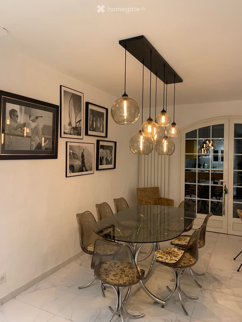 Dining room with a glass table, patterned chairs, pendant lights, and framed pictures on the wall.