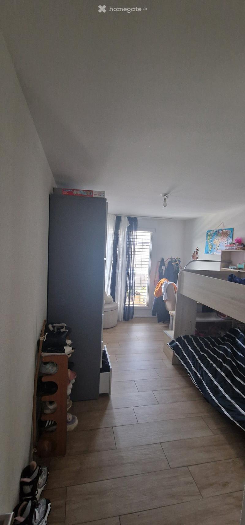 A furnished bedroom with a tiled floor, a bed with striped bedding, a window with curtains, a shelf, a chair, and a shoe rack.
