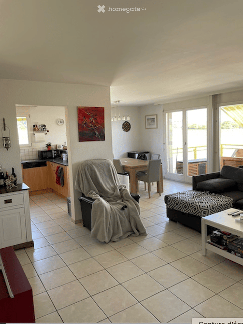 Open-plan living and dining area with tiled flooring, a grey sofa, coffee table, dining table with chairs, and a small kitchen area with a microwave and fridge. A large window with a view to a balcony is visible.