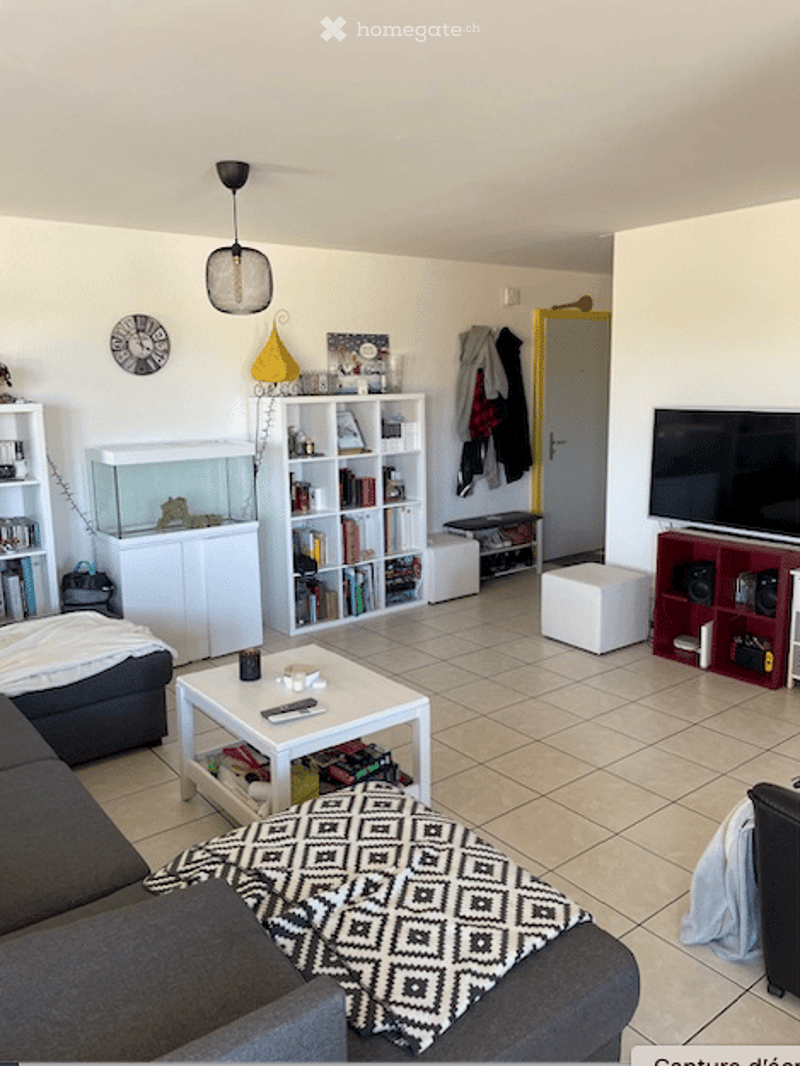 Living room with a grey sofa, coffee table, bookshelf, fish tank, television, and a stool. The room features tiled flooring and a modern aesthetic with a mix of white and dark furniture.