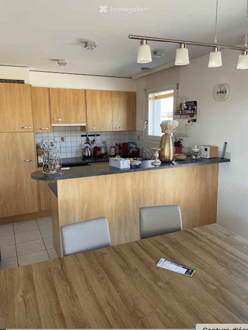 Modern kitchen with light wooden cabinets, a central island, and various appliances including a stove, fridge, microwave, and dishwasher. There is a wooden dining table with chairs and several decorative items on the island.
