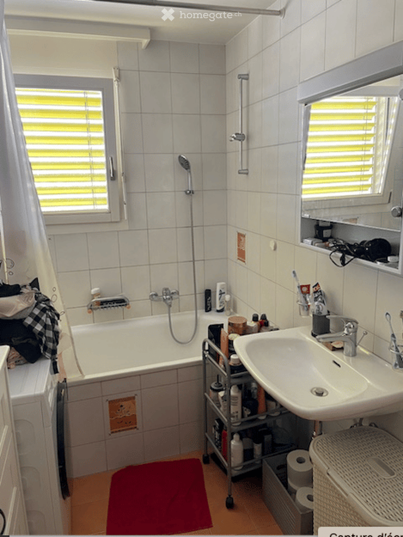 A small bathroom with a white tiled wall, a bathtub, a sink with a mirror, a shower, a towel on the floor, and various toiletries on a small cabinet.