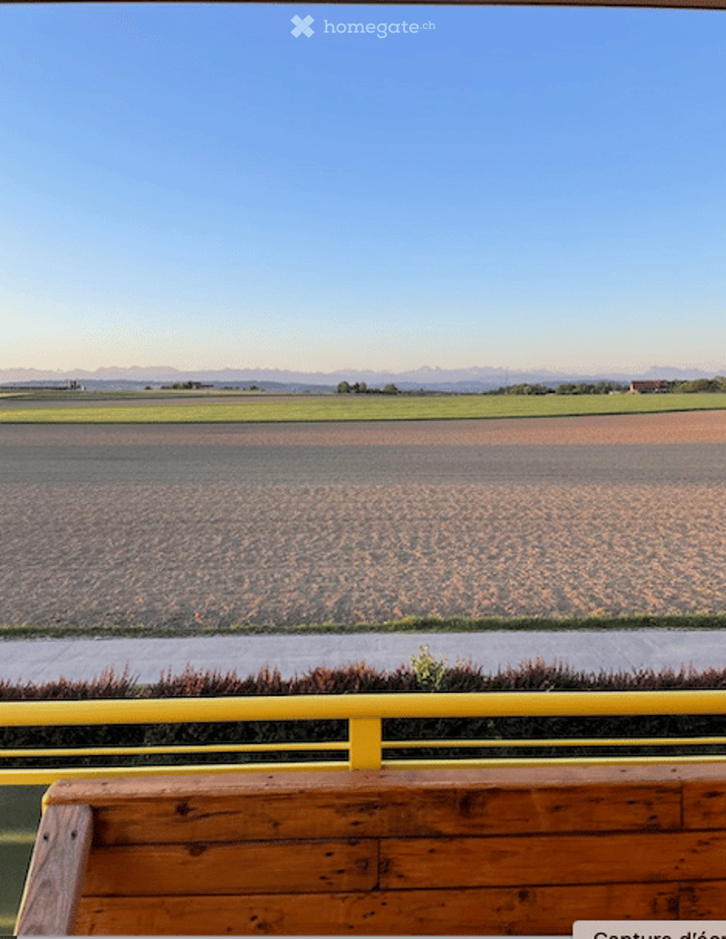 Balcony with yellow railing, wooden bench, overlooking a vast open field and distant mountains under a clear blue sky.