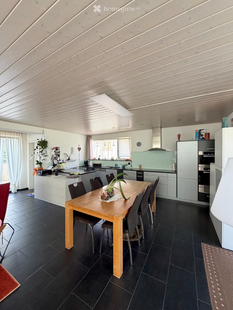 Modern kitchen with a wooden dining table, six chairs, a refrigerator, oven, kitchen island, and various cabinets. The space features black tiled flooring and a sleek, contemporary design with integrated lighting.