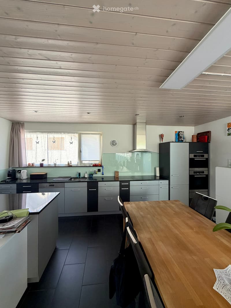 Modern kitchen with grey cabinets, stainless steel appliances, wooden dining table, and black tiled floor.